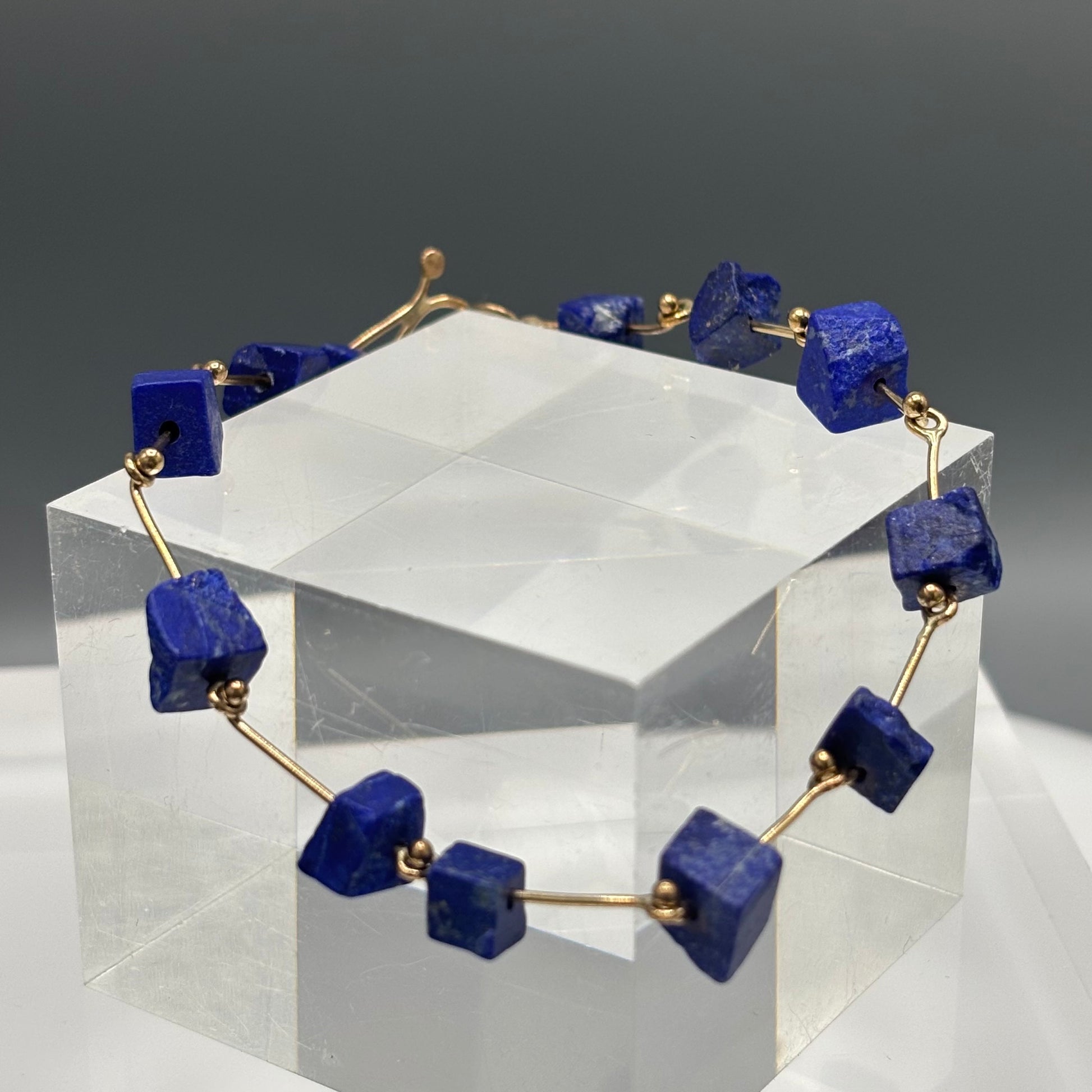 Bracelet with blue square stones and gold accents on a clear stand against a gray background